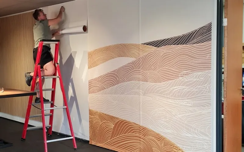 Monteur op een trapje rolt behang met een golvend lijnenpatroon uit over een wand