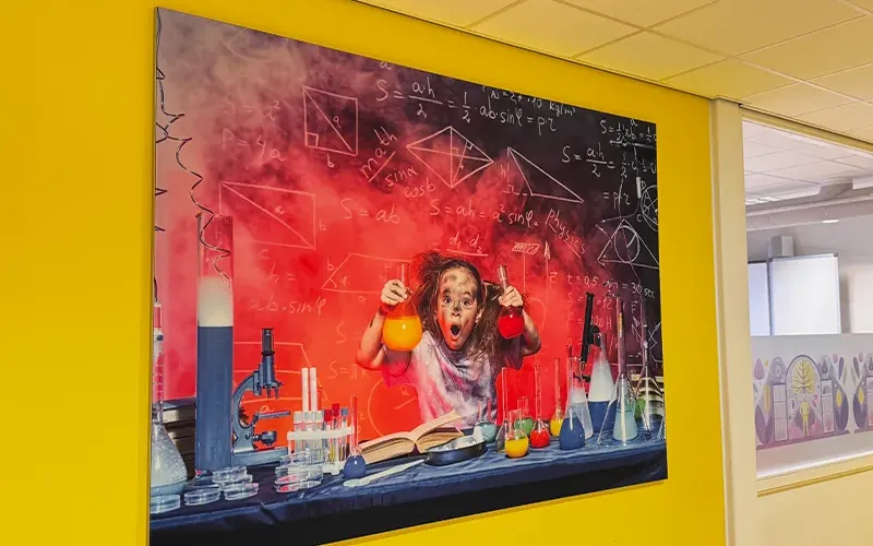Wandpaneel met een print van een scheikundeproefje op een gele muur in een schoolgang