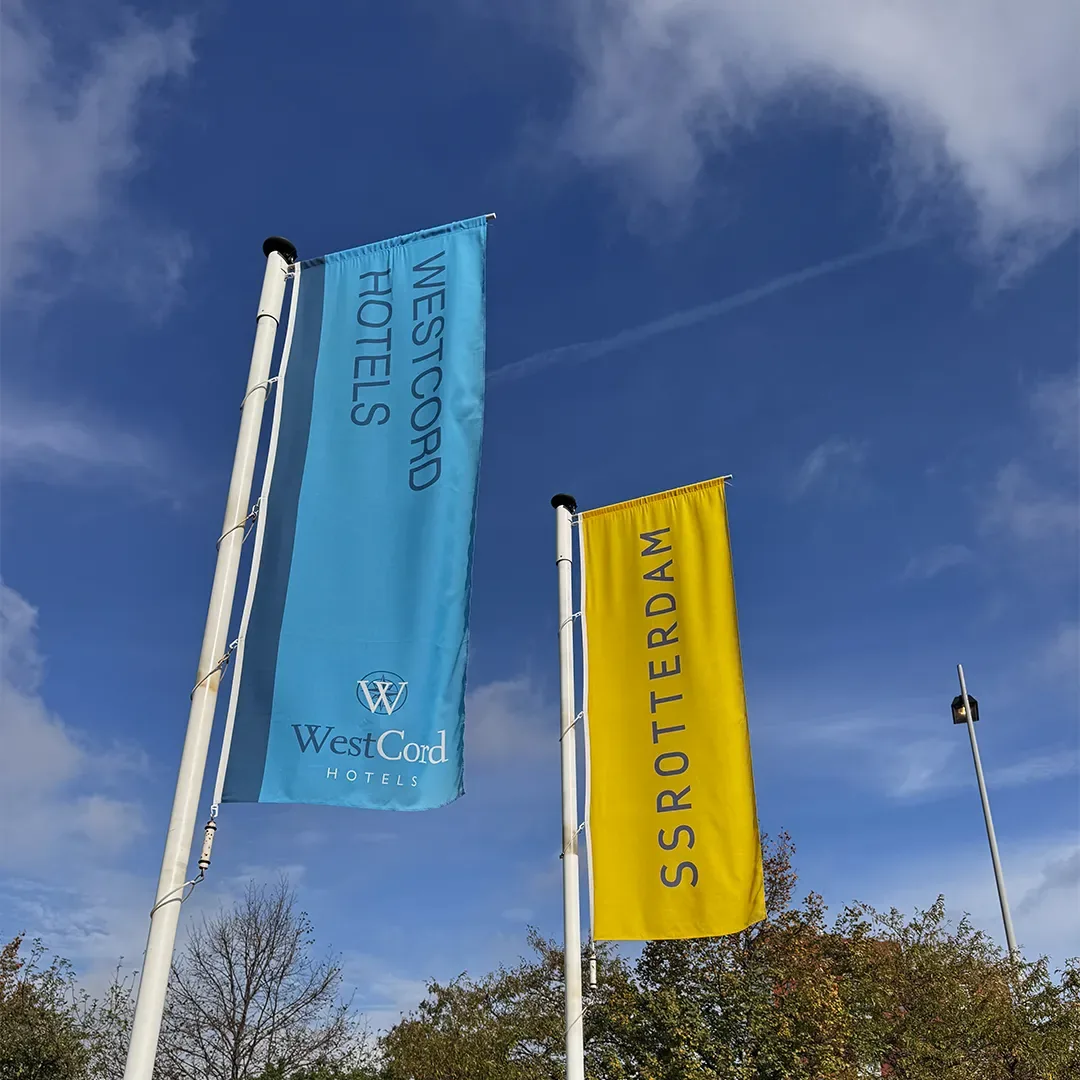 Blauwe WestCord Hotels-vlag en gele SS Rotterdam-vlag aan twee masten