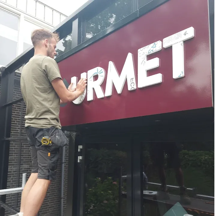 Monteur drukt de witte letters op de rode Gourmet-lichtbak, beschermfolie zit er nog op