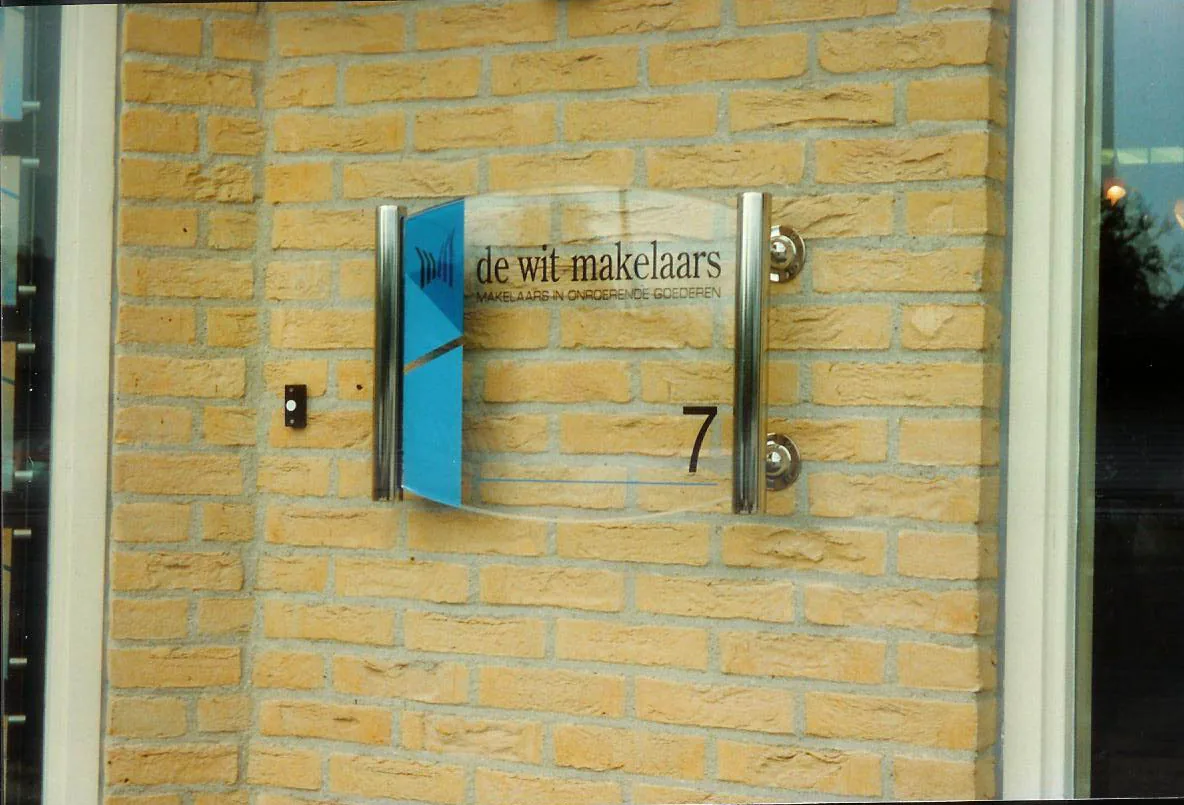 Glazen naambord van De Wit Makelaars op afstandhouders aan een gele gevel
