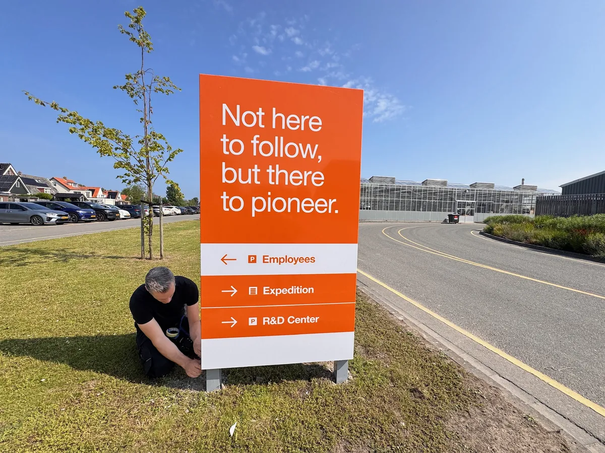 Monteur zet een oranje bord met de tekst Not here to follow, but there to pioneer en richtingpijlen in het gras