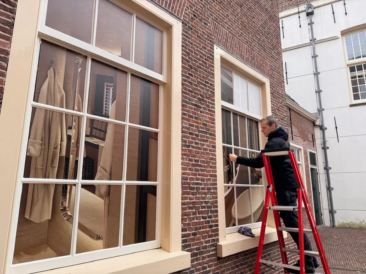 Raam met een print van een hotelkamer, een monteur op een trap ernaast