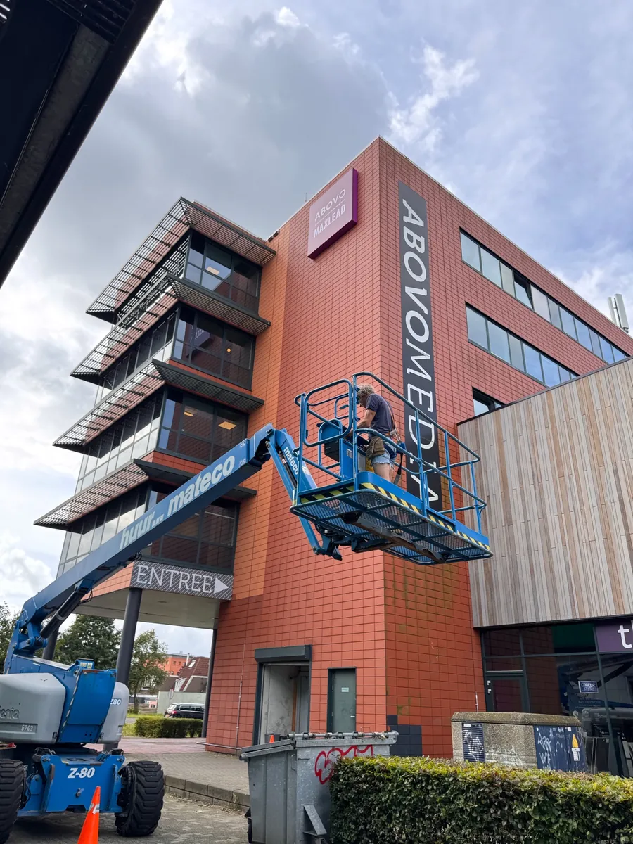 Hoogwerker bij de verticale naam ABOVOMEDIA op het rode kantoorpand