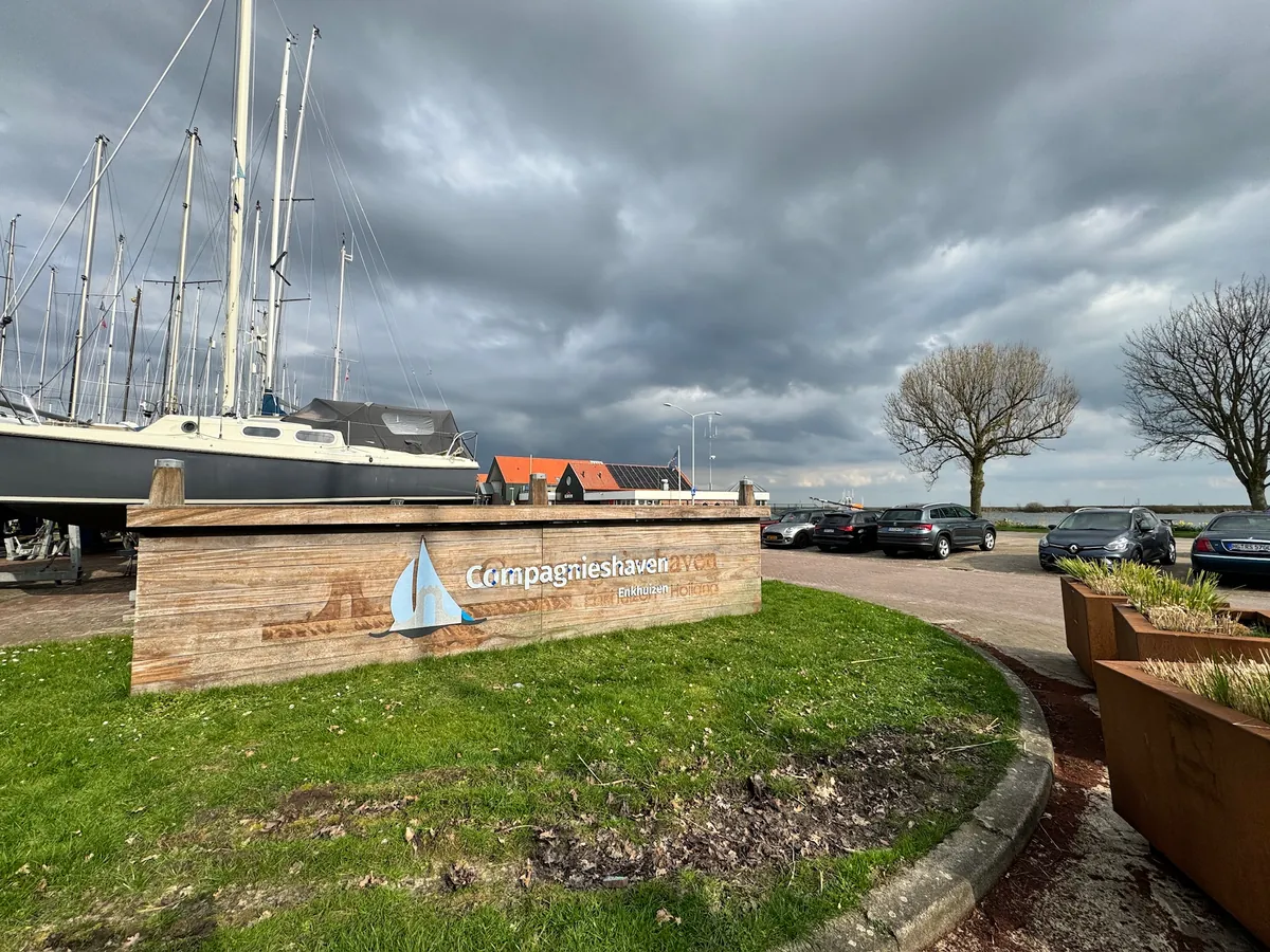 Houten bord met het Compagnieshaven-logo in het gras bij de haven, onder een bewolkte lucht