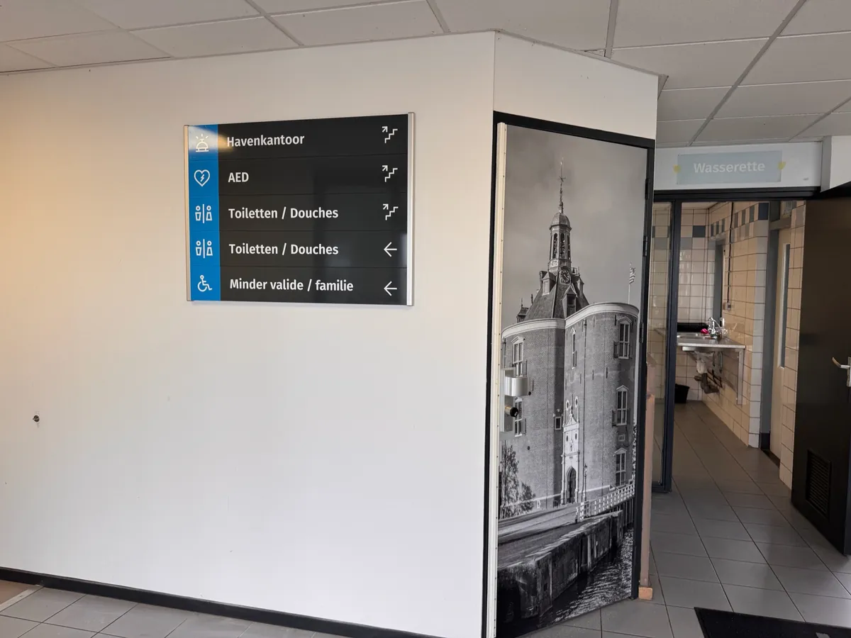 Wandbord met pijlen naar havenkantoor, AED en toiletten, naast een zwart-witprint van een toren