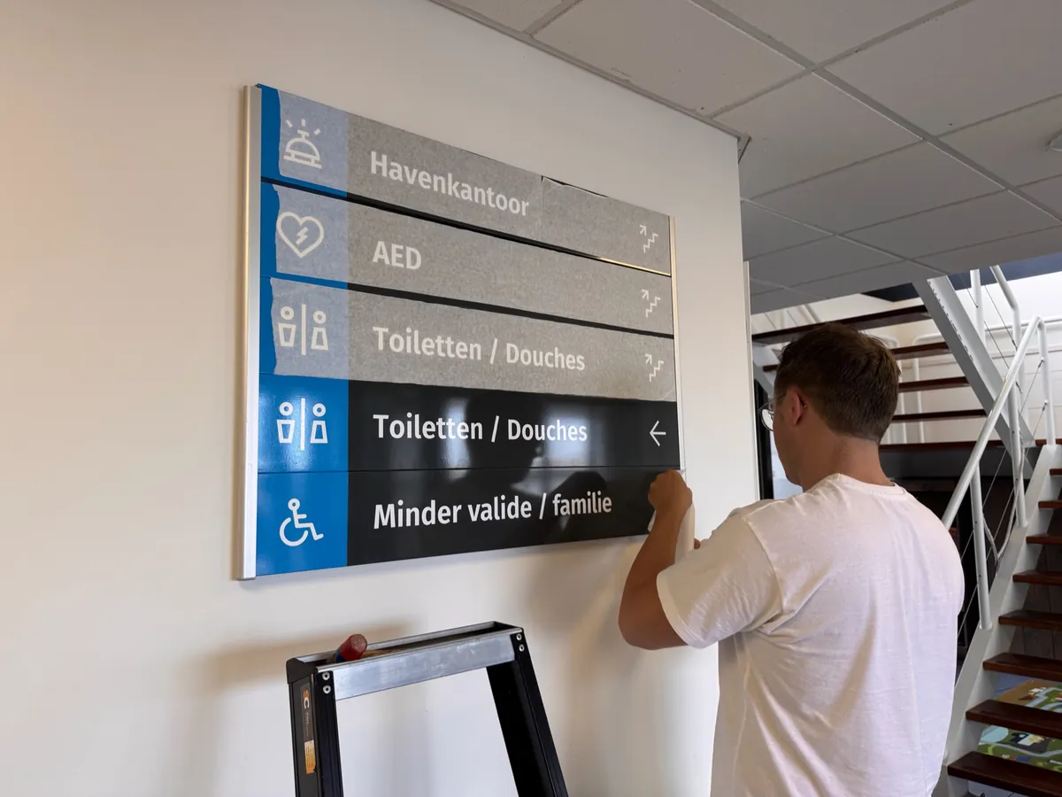 Monteur hangt het wandbord met pijlen naar havenkantoor, AED en toiletten