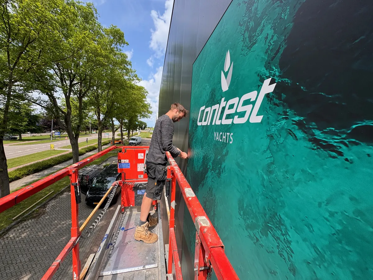 Monteur op een hoogwerker werkt aan het groene gevelbeeld met het Contest Yachts-logo