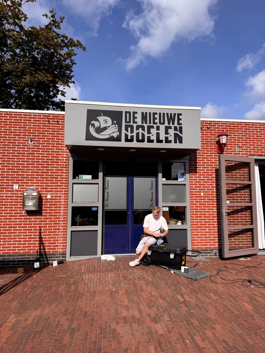 Bakstenen gevel met het naambord DE NIEUWE DOELEN boven de entree, een monteur op de stoep