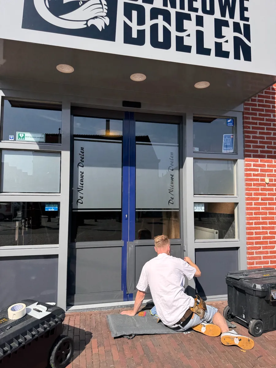 Monteur zit op de stoep en brengt matte raamfolie met de tekst De Nieuwe Doelen aan op de deuren