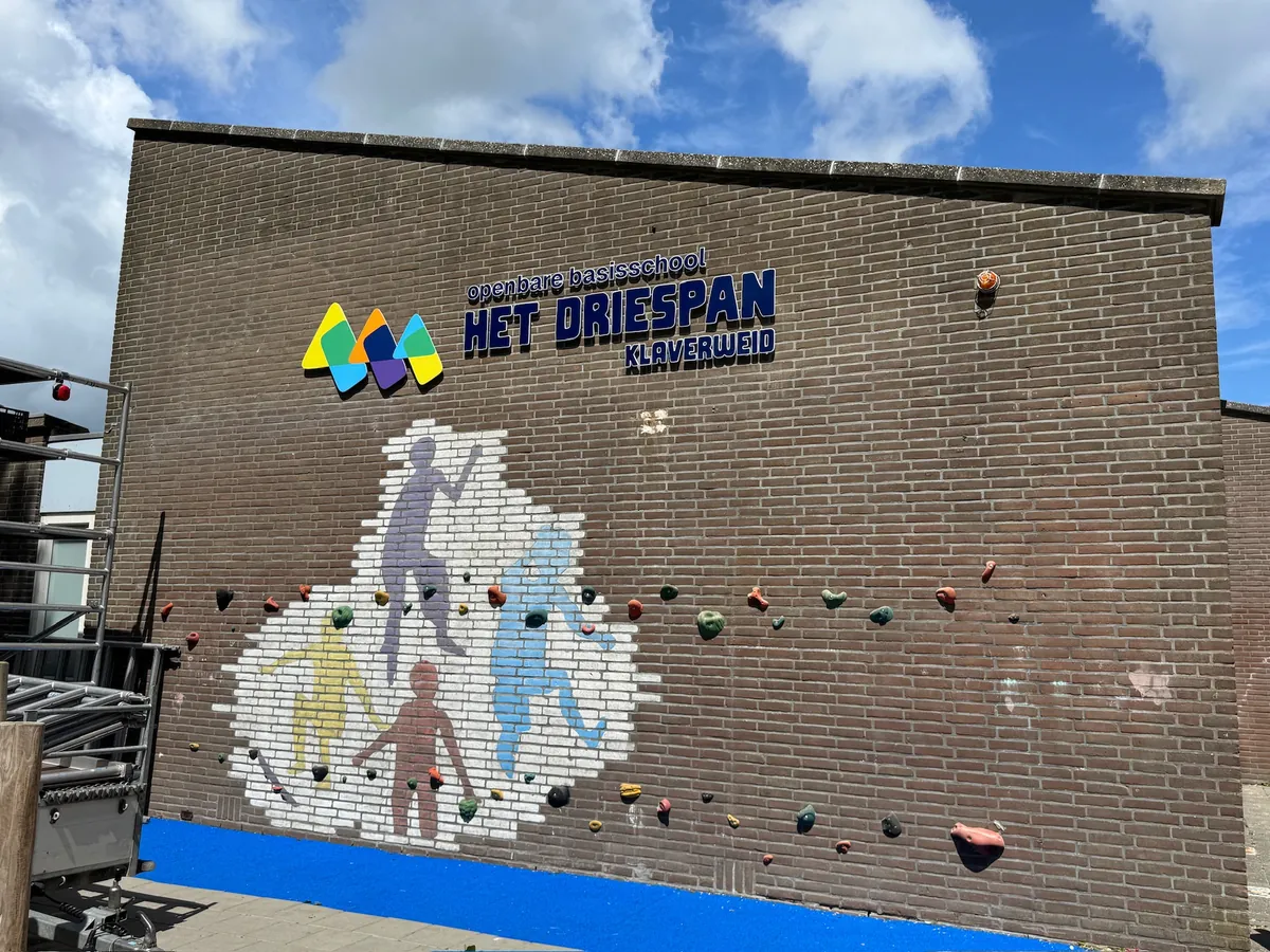 Bakstenen muur met het schoollogo en een wit vlak met figuren en gekleurde klimgrepen