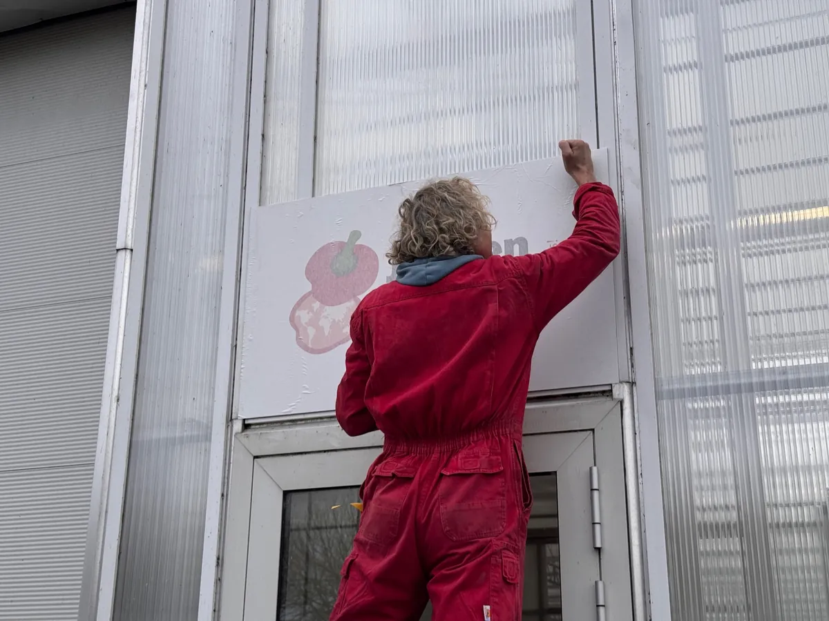 Monteur in rood werkpak hangt een bord op boven een deur van de kas