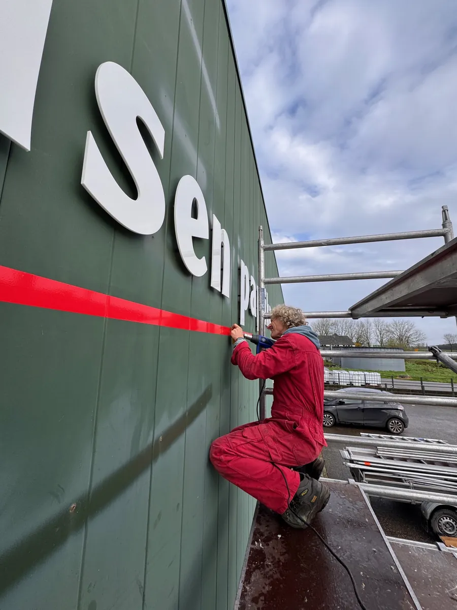 Monteur in rood werkpak zet de witte letters van Jansen paprika langs een rode lijn op de groene gevel