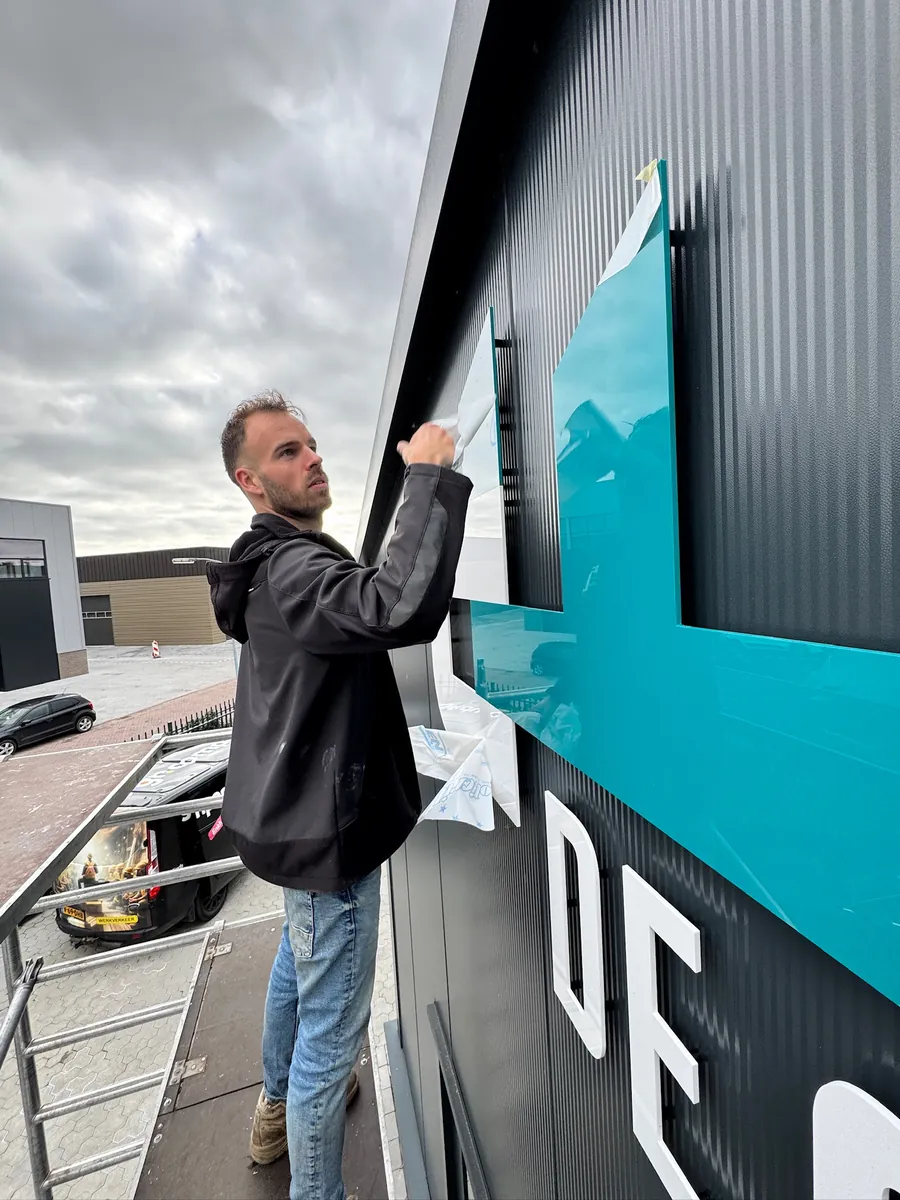 Monteur op een hoogwerker plaatst het turquoise logodeel op de gevel