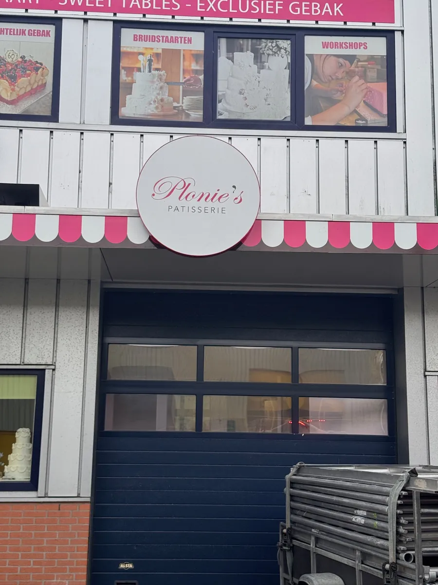 Rond bord van Plonie's Patisserie boven de roze-witte zonwering en roldeur