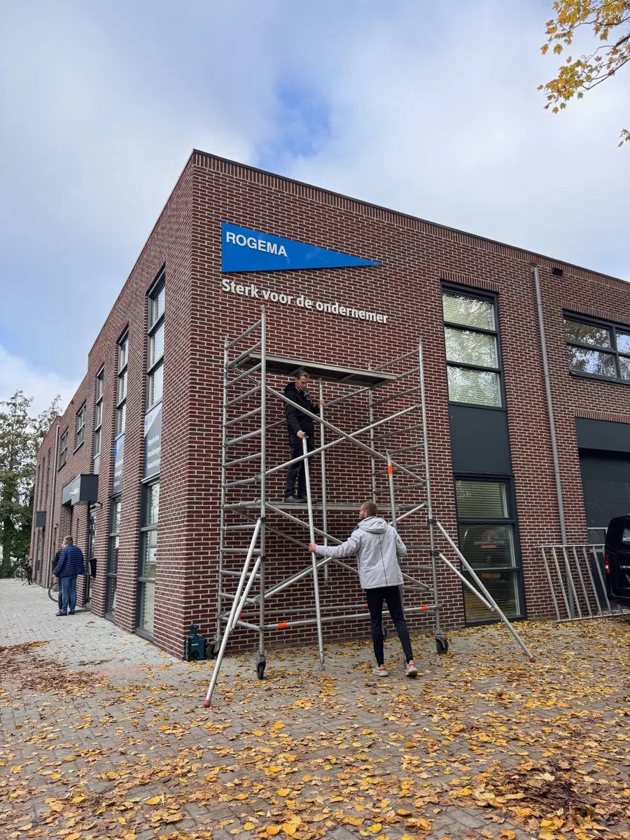 Monteurs bij een rolsteiger tegen de bakstenen gevel met het Rogema-logo
