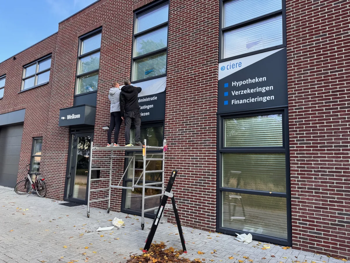 Twee monteurs op een steiger brengen raambelettering aan boven de entree