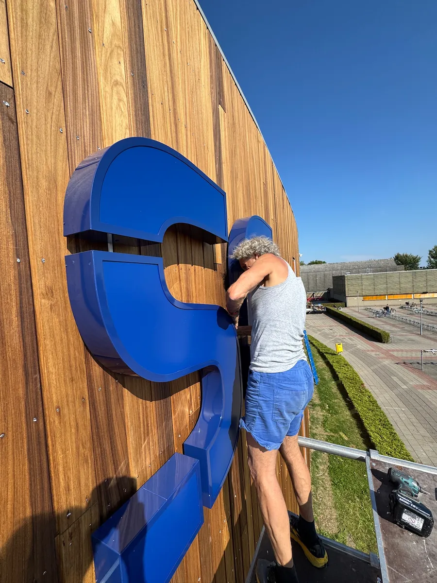Monteur houdt een grote blauwe letter vast tegen de houten gevel