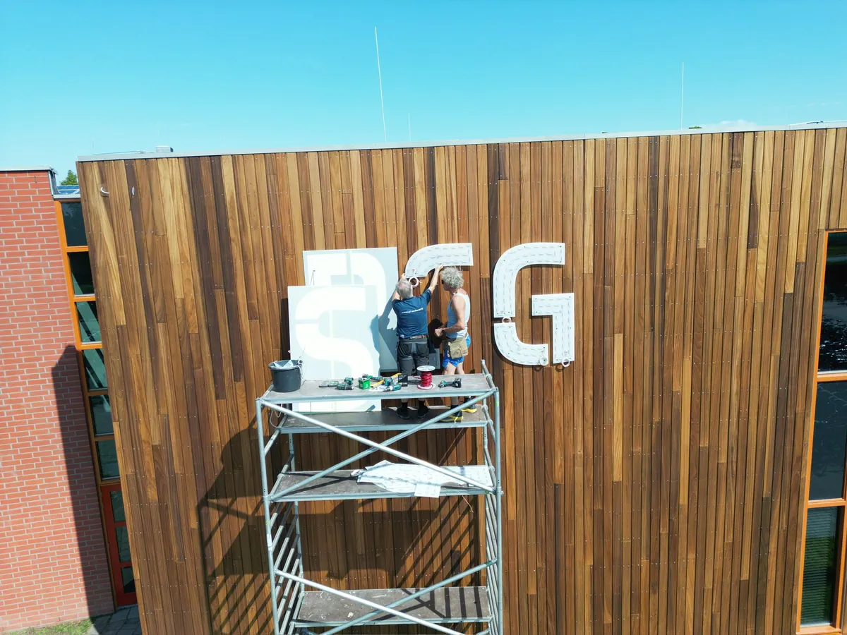Twee monteurs op een rolsteiger zetten de grote letters RSG op de houten gevel
