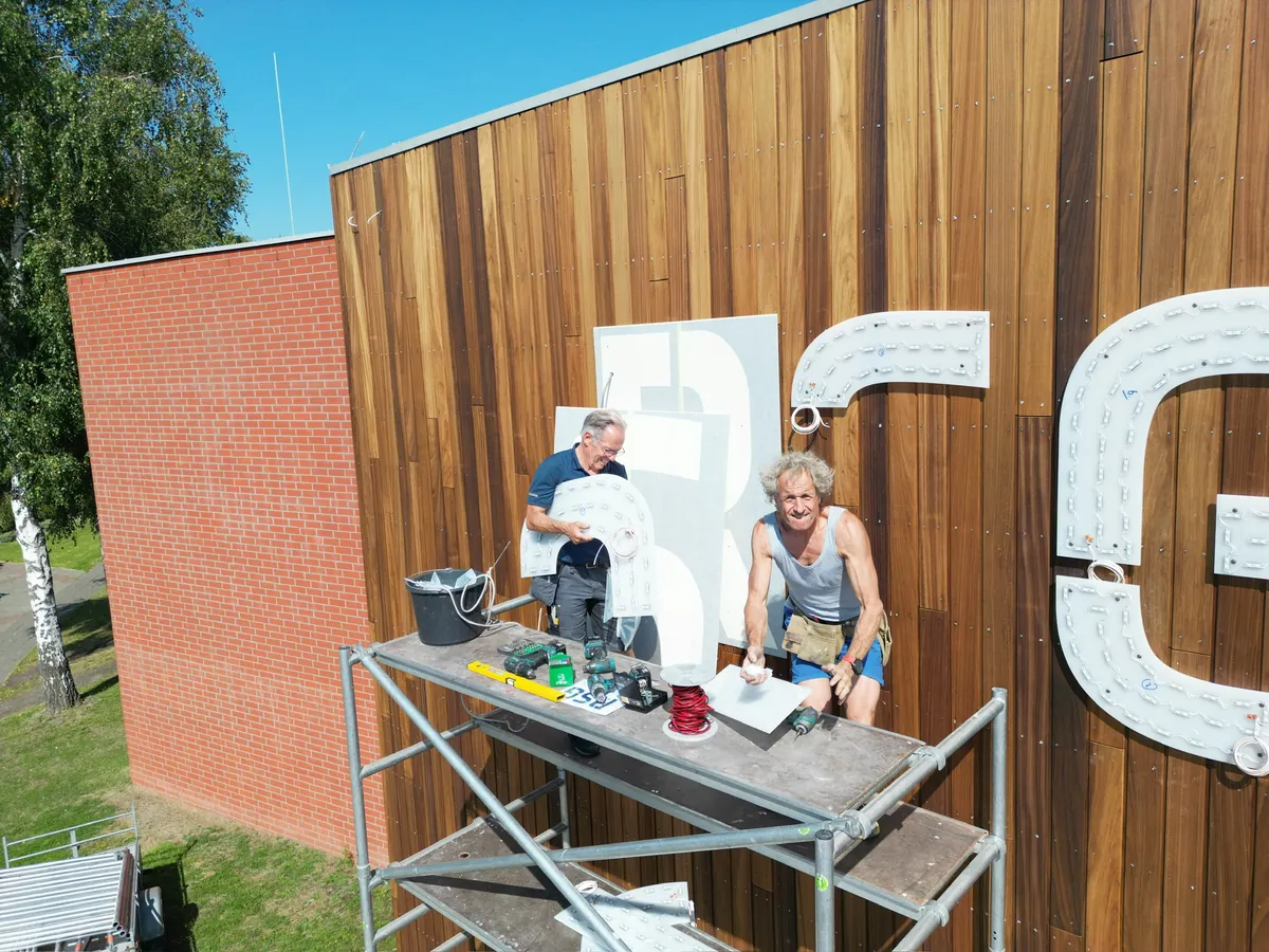 Twee monteurs op de steiger met een witte letter R, naast de G op de houten gevel