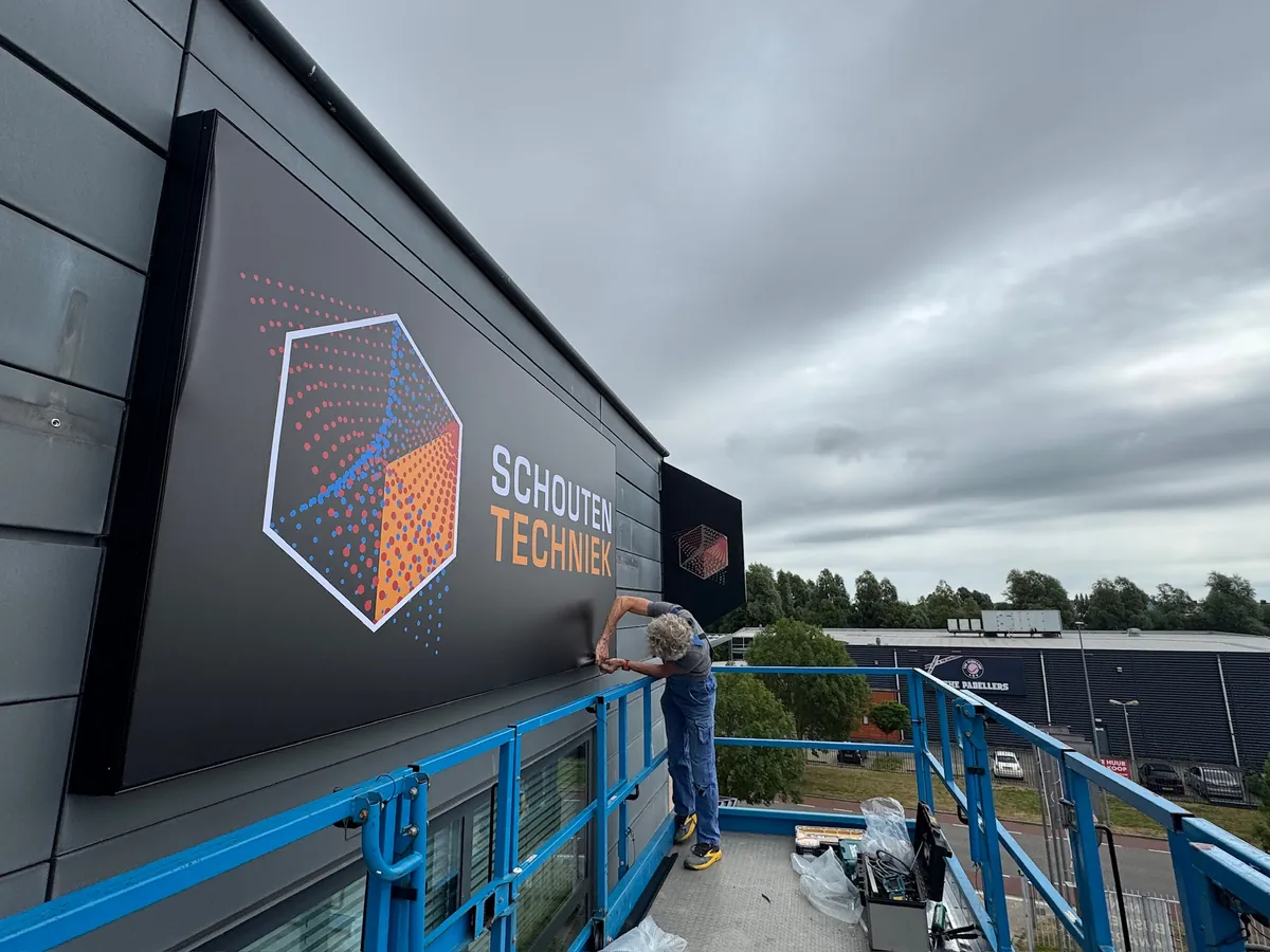 Groot donker bord met het Schouten Techniek-logo op de dakrand, met een monteur op de hoogwerker