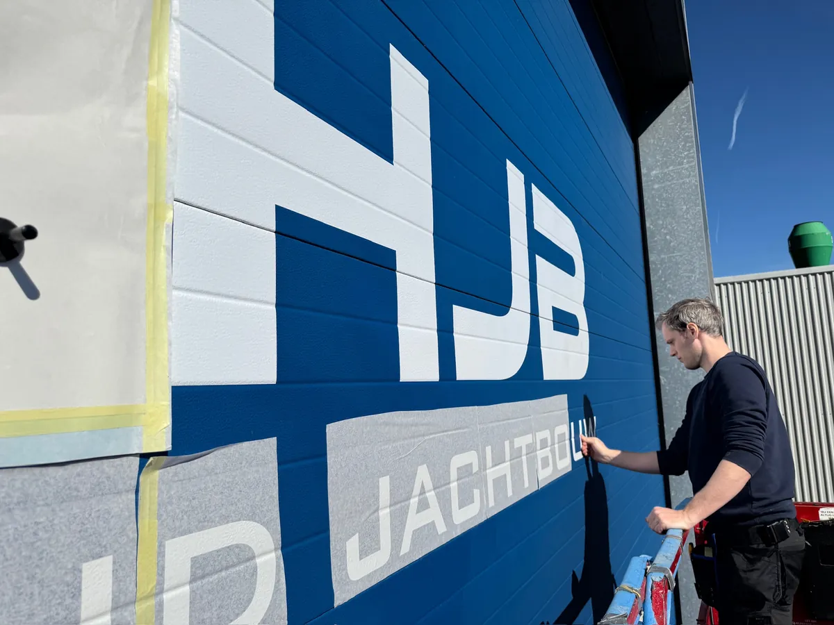 Monteur brengt de witte letters van HJB Jachtbouw aan op de blauwe roldeur