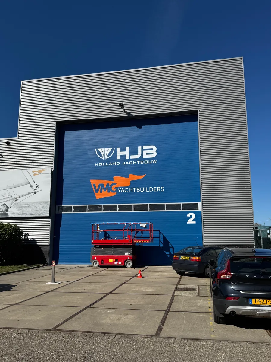 Blauwe loodsdeur met de logo's van HJB Holland Jachtbouw en VMG Yachtbuilders
