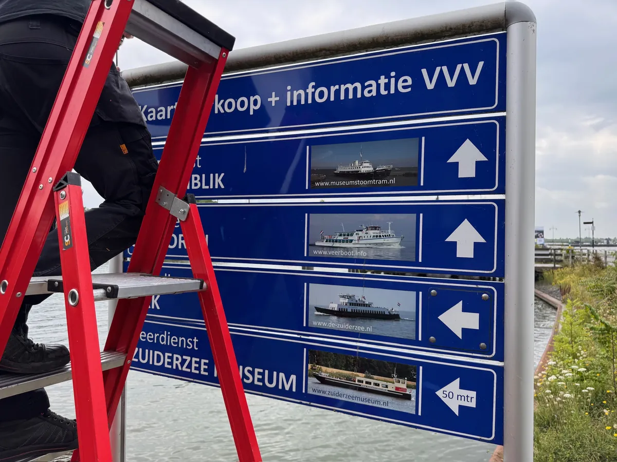 Monteur op een rood trapje bij een blauw richtingbord met pijlen naar de VVV en het Zuiderzeemuseum