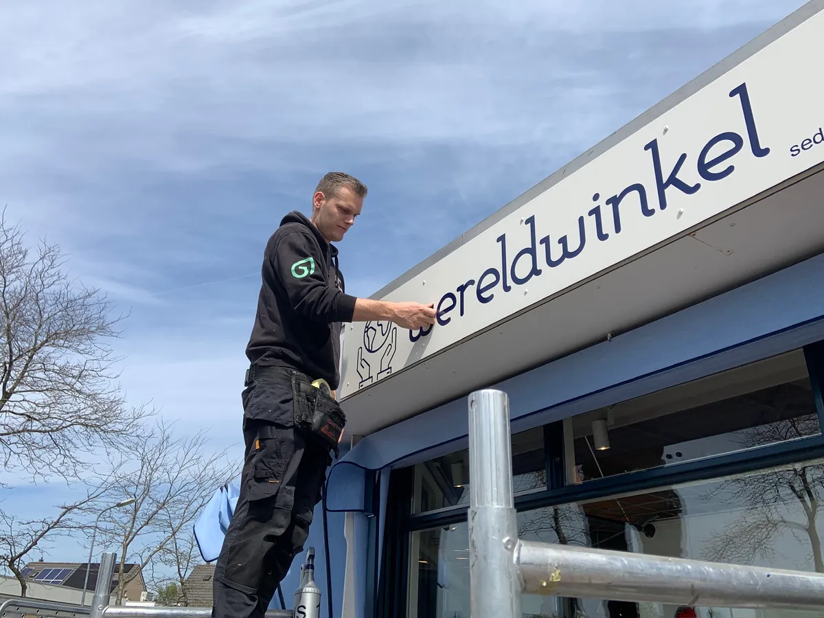 Monteur brengt de naam wereldwinkel aan op de witte gevelrand