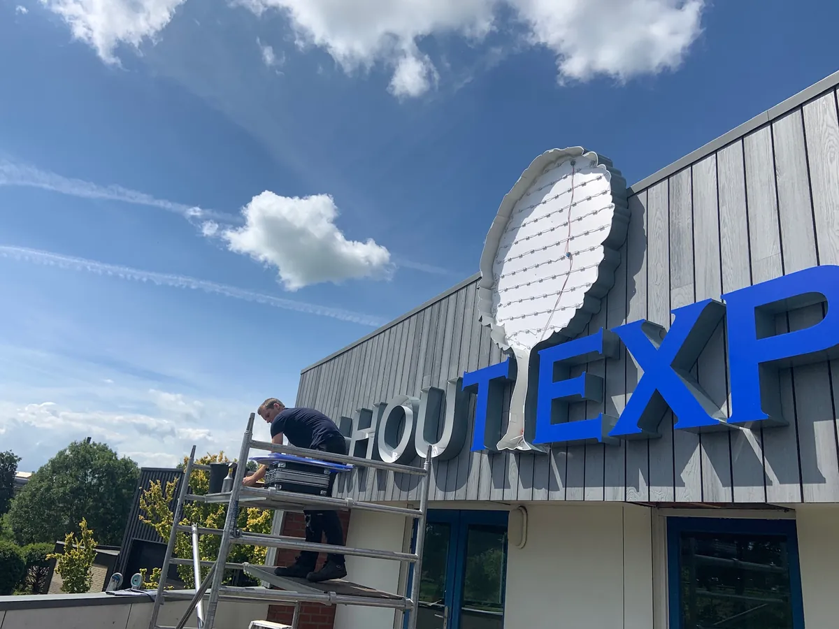 Monteur op een steiger plaatst blauwe letters van Hout Expert op de grijze gevel