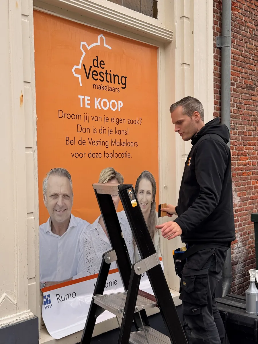 Monteur op een trapje brengt een oranje TE KOOP-raambeeld van de Vesting makelaars aan
