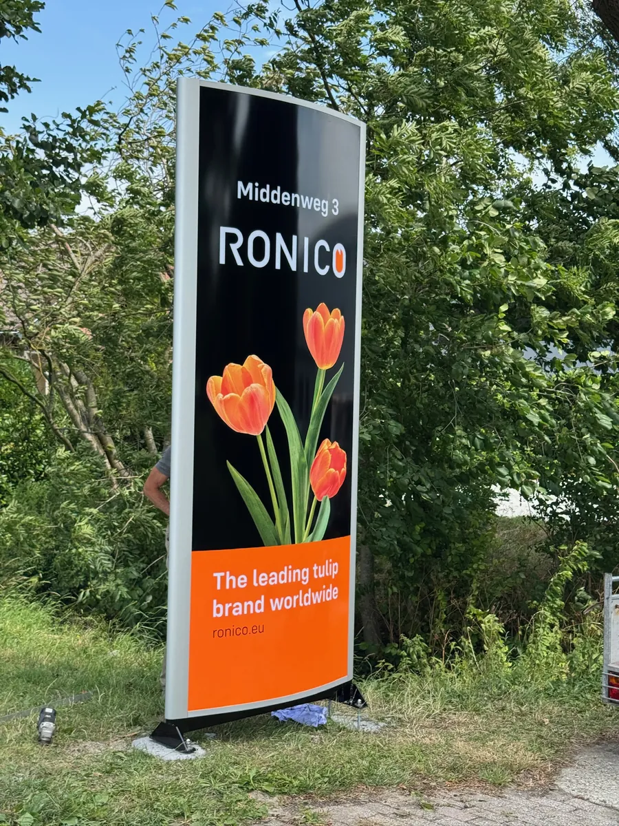 Zwarte reclamezuil van Ronico met oranje tulpen, langs de oprit