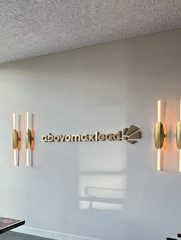 Gouden RVS-letters met het logo van Abovomaxlead op een lichte binnenmuur, tussen twee wandlampen