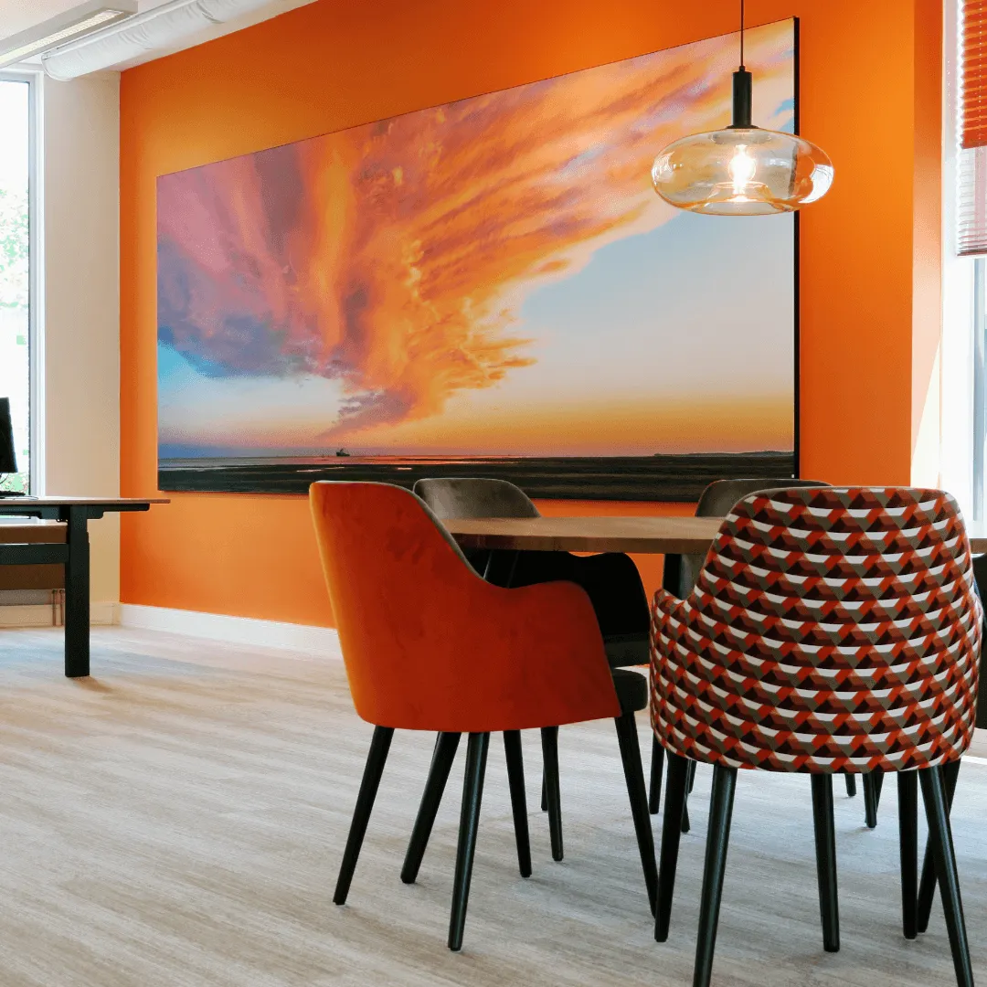Panoramisch wanddoek met een oranje wolkenlucht boven het wad, aan een oranje kantoorwand boven de vergadertafel