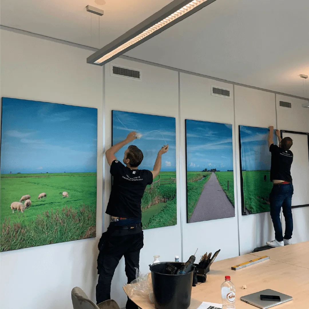 Twee monteurs van Sign Company hangen een reeks doeken met polderfoto's op in een vergaderruimte